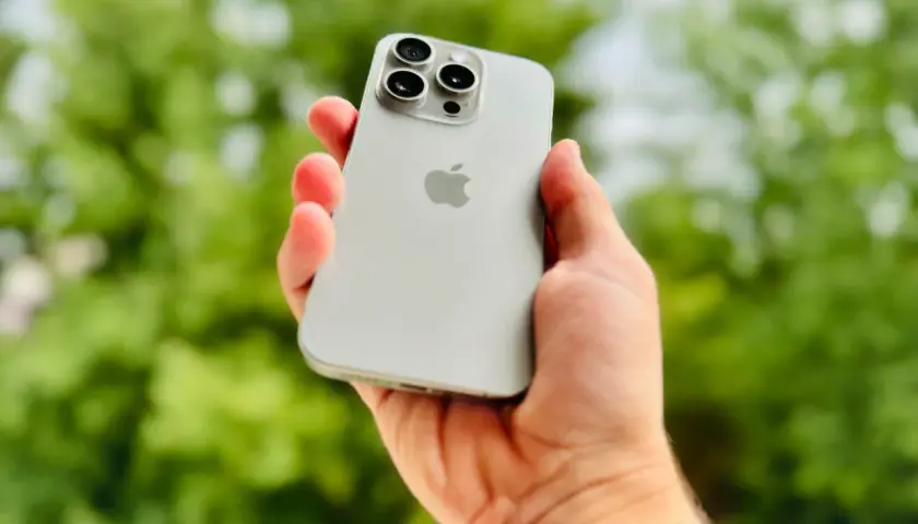 Dłoń trzyma srebrny iPhone 15 Pro Max, prezentując jego potężny aparat.