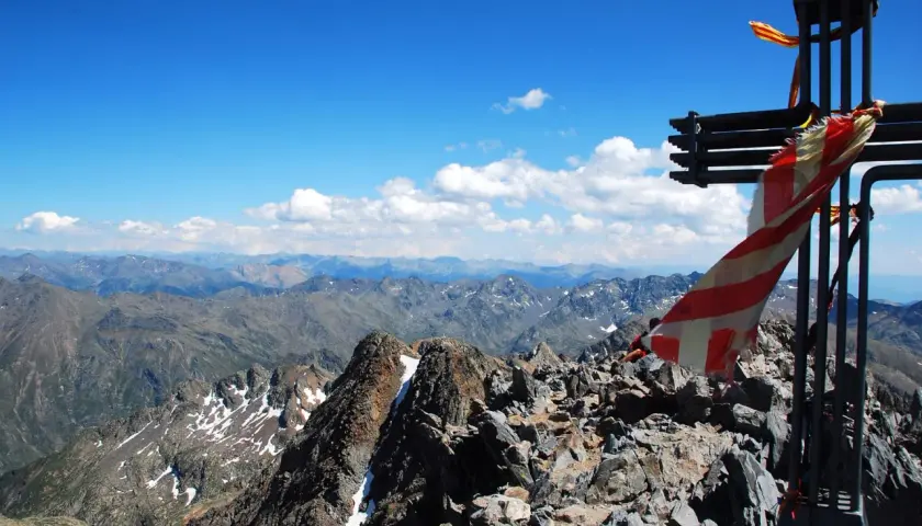 Cruz metálica en la cima de una pica d'estats, con bandera ondeando y un paisaje montañoso al fondo.