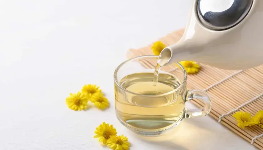 Tè giallo dorato versato da una teiera in una tazza trasparente, circondato da piccoli crisantemi fiori gialli.