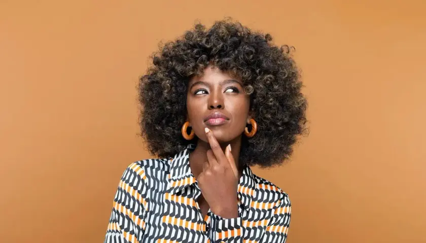 Mujer pensativa con cabello rizado y coloración semipermanente, luciendo una blusa estampada y aretes circulares.