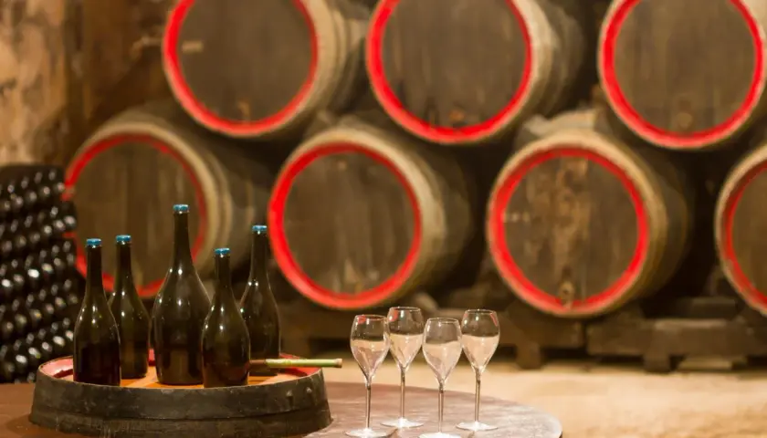 Bouteilles de champagne et verres sur un tonneau, prêts pour une visite de cave. Au fond, des fûts de chêne empilés.