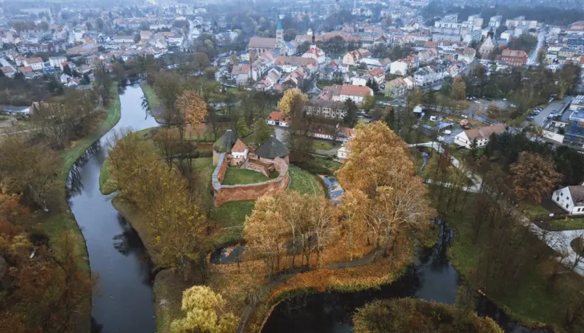 Zamek międzyrzecz otoczony jesiennymi drzewami i wodą, z miastem w tle.
