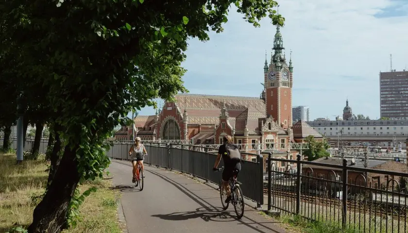 Dwie osoby na rowerach jadą ścieżką obok torów kolejowych. W tle piękny budynek dworca z wieżą zegarową. To idealne miejsce przyjazne rowerzystom.