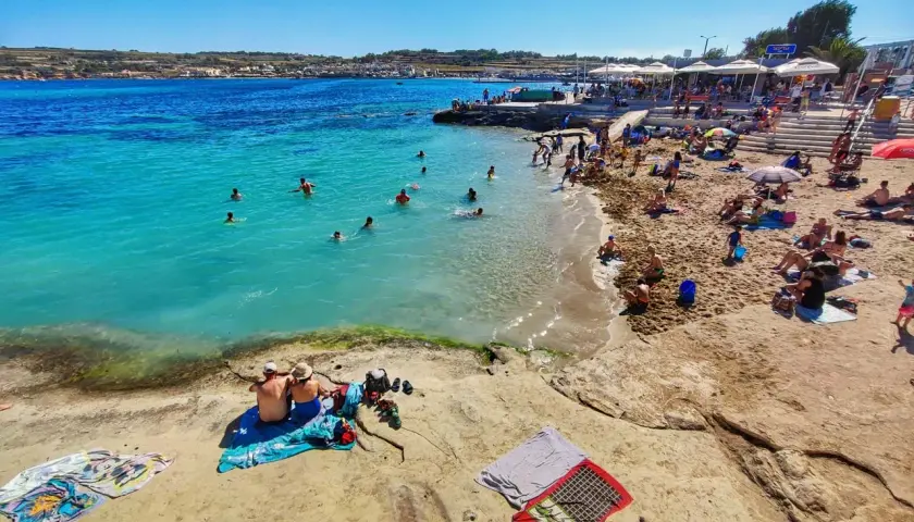 Słoneczna plaża na Malcie z turystami kąpiącymi się w turkusowym morzu i odpoczywającymi na piasku.