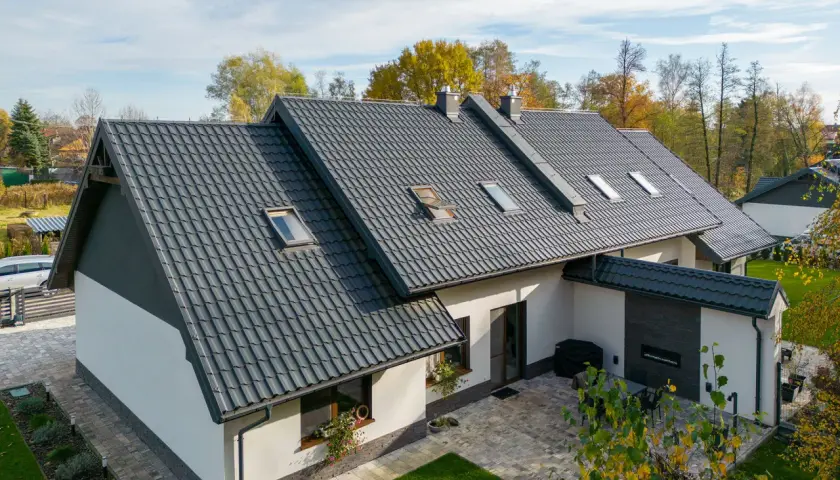 Nowoczesny dom z białymi ścianami i ciemnym, eleganckim dachem z dachówki ceramicznej. Wokół zieleń i jesienne drzewa.