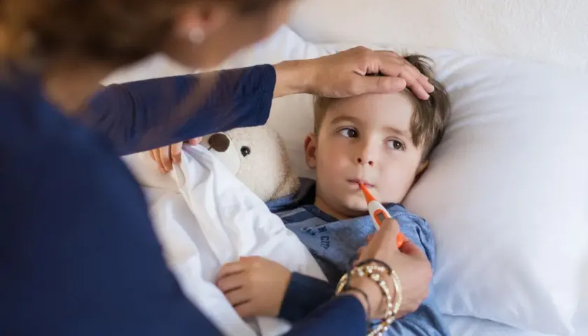 Chłopiec leży w łóżku z termometrem w ustach, mama sprawdza mu czoło. Widać, że jest w stanie podgorączkowym.