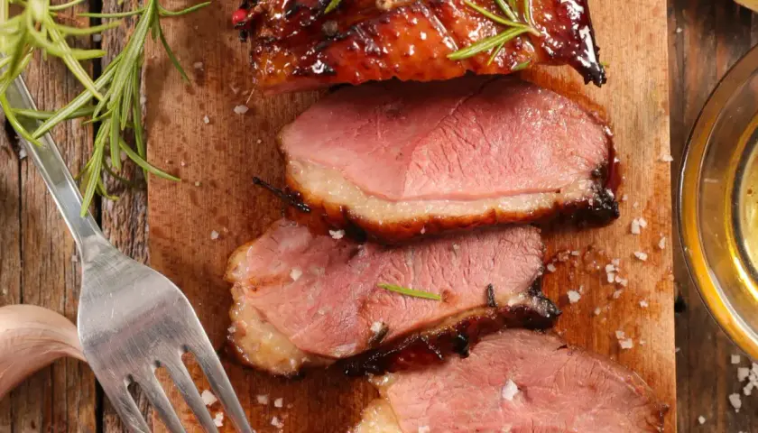 Magret de canard rosé, tranché sur une planche en bois, garni de romarin. Une fourchette et une gousse d'ail sont à proximité. Un délice pour accompagner un bon vin.
