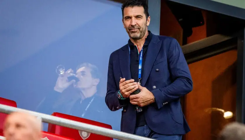 Gianluigi Buffon, legendarny włoski bramkarz, w eleganckim granatowym garniturze, obserwuje mecz z trybun.