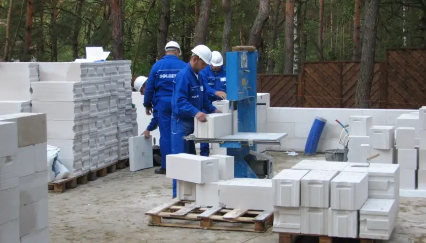 Na budowie pracownicy w niebieskich kombinezonach i kaskach układają bloczki betonowe. Widać kilka palet z bloczkami, gotowych do użycia.