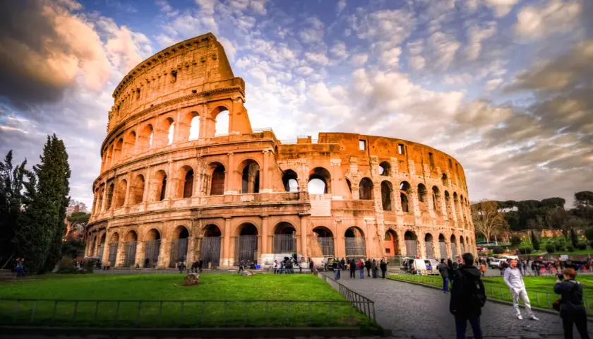 Il Colosseo, un'icona di cosa visitare a Roma, si erge maestoso sotto un cielo drammatico. Turisti passeggiano sul selciato, ammirando l'antica grandezza.