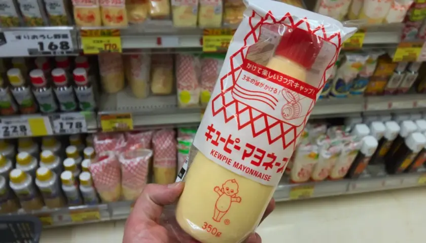 A hand holds a large squeeze bottle of Kewpie mayonnaise. The packaging features the iconic Kewpie doll. The question "how long does Kewpie mayo last?" is on my mind as I shop.