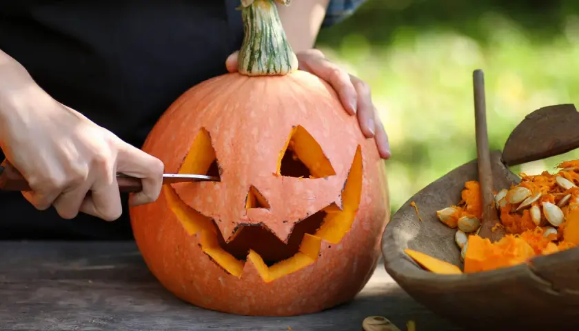 Händer skär ett ansikte i en pumpa för en klassisk pumpasminkning. Bredvid ligger en skål med pumpafrön och fruktkött.