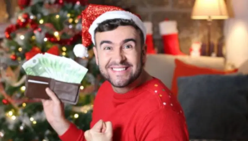 Hombre con gorro de Papá Noel muestra billetera llena de billetes, como si soñara que le toca la lotería en Navidad.