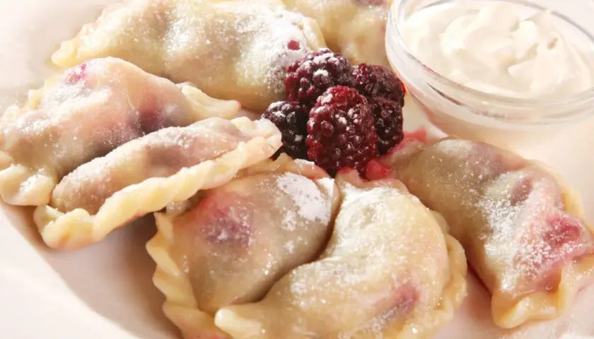 Pyszne pierogi z malinami, posypane cukrem pudrem, podane z kwaśną śmietaną i świeżymi owocami leśnymi.
