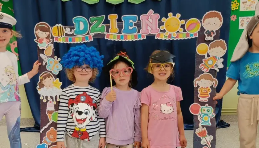 Dzieci w przedszkolu w strojach i z rekwizytami pozują do zdjęcia na tle dekoracji z napisem "Dzień Dziecka".