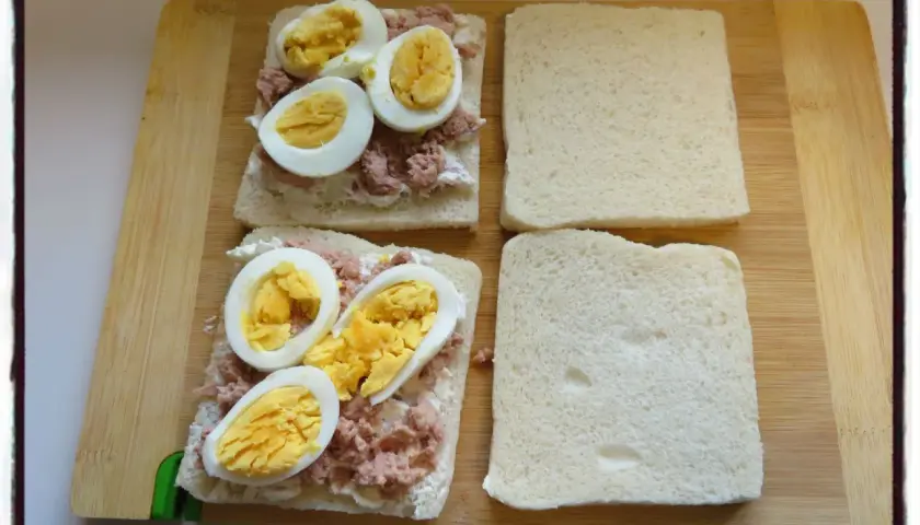 Deux sandwichs oeuf dur sont en préparation sur une planche en bois. Le thon émietté et la mayonnaise sont déjà étalés sur le pain.