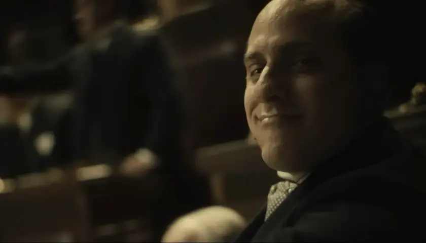 Un uomo con un sorriso furbo sembra rompere la quarta parete, guardando direttamente lo spettatore.