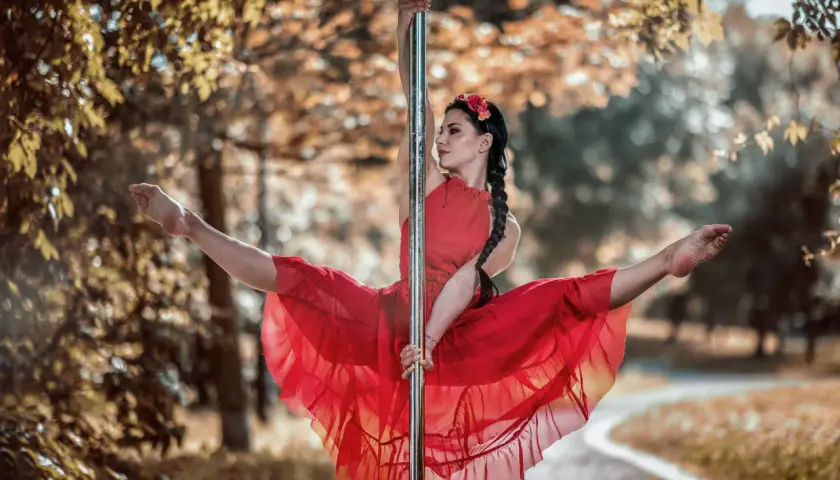 Kobieta w czerwonej sukience wykonuje figurę na słupie, jak podczas kursu pole dance. Jej nogi są rozłożone w szpagacie, a włosy splecione w warkocz.