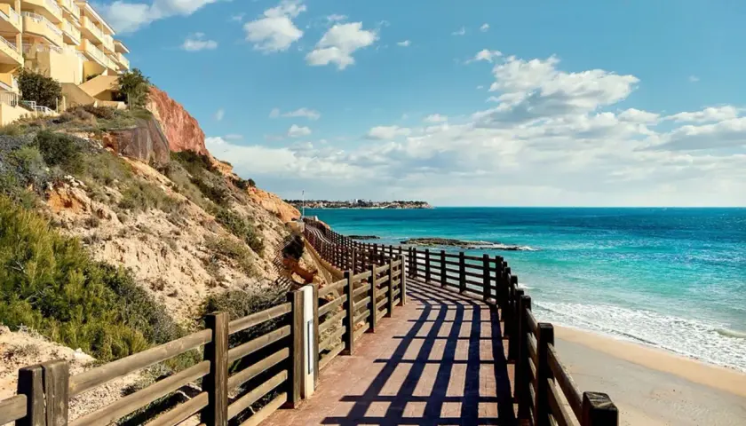 Paseo de madera con vistas a las mejores playas de Cabo Roig. Mar turquesa, arena dorada y un edificio amarillo en la ladera.