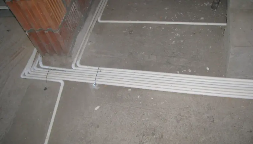 Instalacja elektryczna w podłodze, przygotowana zgodnie z przepisami. Białe rury układają się wzdłuż betonowej posadzki, obok ceglanej ściany.