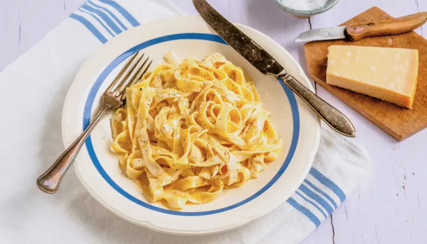 Ein Teller cremiger Fettuccine, bestreut mit schwarzem Pfeffer, daneben ein Stück Parmesan und Salz.