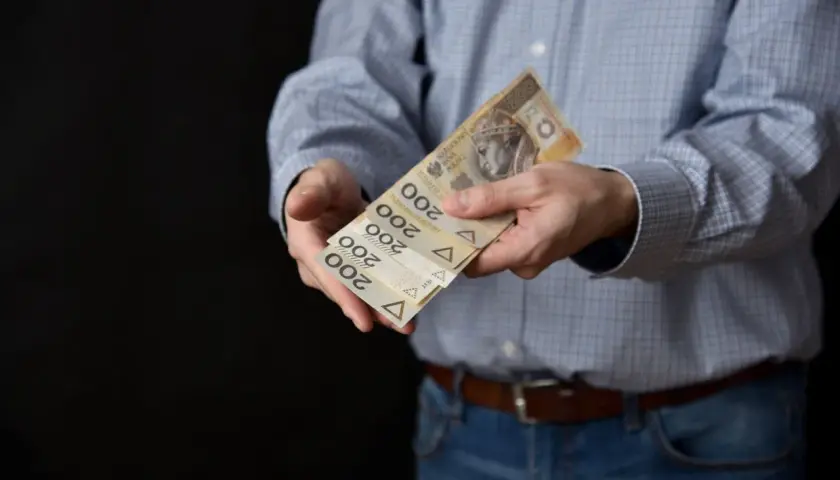 Mężczyzna w koszuli i jeansach podaje kilka banknotów po 200 zł. Może to być symbol zapłaty, darowizny lub cesji.