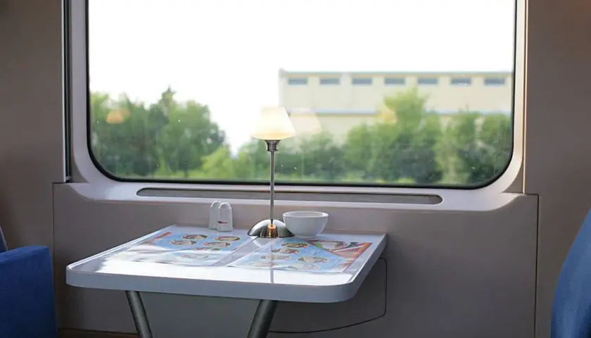 Stolik w wagonie restauracyjnym z lampką, solniczką i menu. Za oknem zieleń i budynek.