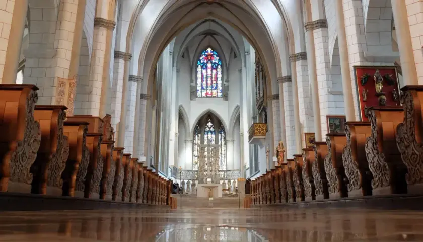 Blick durch das Kirchenschiff des Augsburger Doms mit seinen kunstvoll geschnitzten Holzbänken und dem farbenprächtigen Chorfenster.