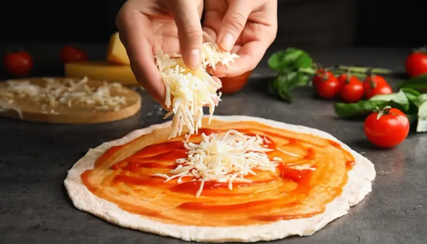 Hände streuen geriebenen Gouda auf eine mit Tomatensauce bestrichene Pizza. Tomaten und Basilikum liegen daneben.