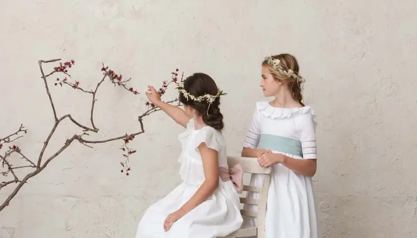 Dos niñas con peinados de comunión con diadema floral, una sentada tocando ramas, la otra de pie.