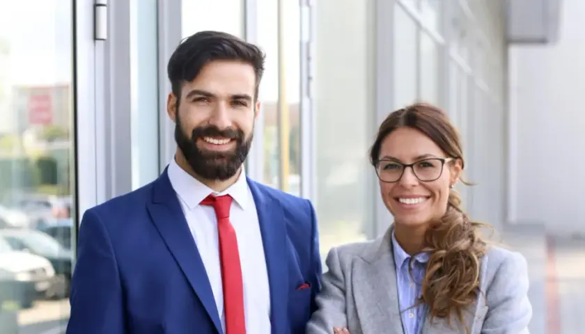 Smiling professionals, a man in a blue suit and red tie, and a woman in a grey blazer, stand outside. They're ready to pursue online MPA programs no GRE.