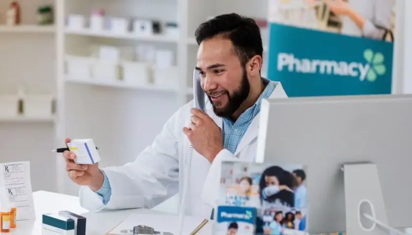 Uśmiechnięty farmaceuta rozmawia przez telefon, trzymając pudełko leku. Zastanawia się, ile zarabia farmaceuta, pracując w aptece.