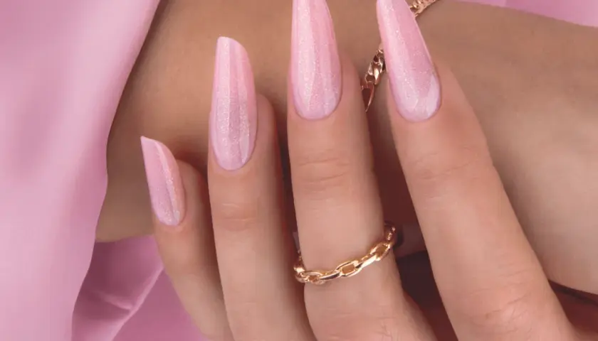 Delikatny, różowy manicure hybrydowy z subtelnymi drobinkami brokatu. Długie paznokcie w kształcie migdała, ozdobione złotą biżuterią.