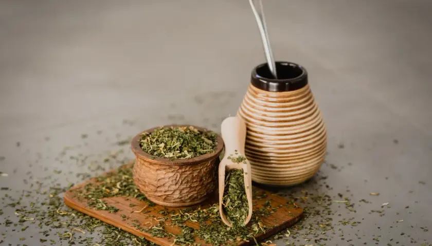 Drewniana miseczka i łopatka pełne suszonej yerba mate, obok ceramiczny naczynie z bombillą. Napar z yerba mate wspiera wątrobę.