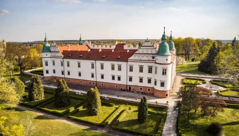 Zamek w Baranowie Sandomierskim, biała rezydencja z czerwonym dachem i zielonymi wieżyczkami, otoczona zadbanym ogrodem.