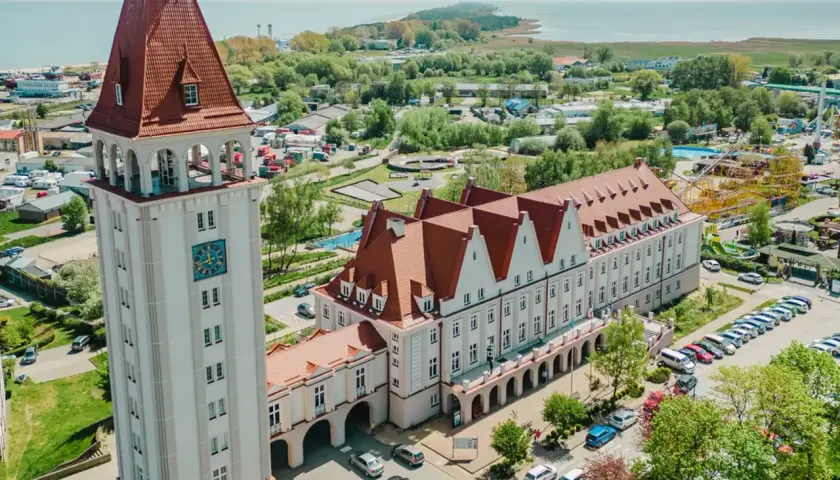 Wieża zegarowa i długi budynek z czerwonym dachem, przypominający dom rybaka, stoją obok siebie. W tle widać morze i park rozrywki.