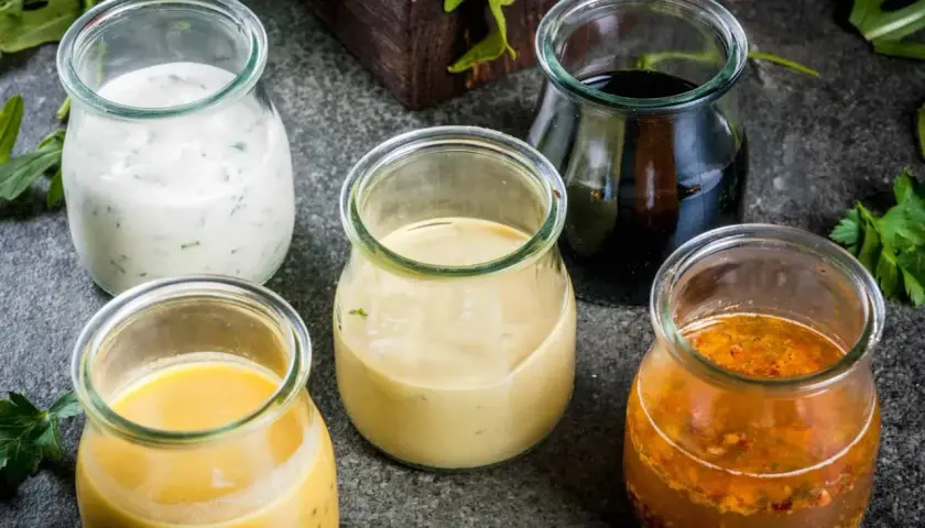 Fem små glasburkar med olika dressingar till sallad: en vit med örter, en gul, en ljusbrun, en mörk och en med frön och kryddor.
