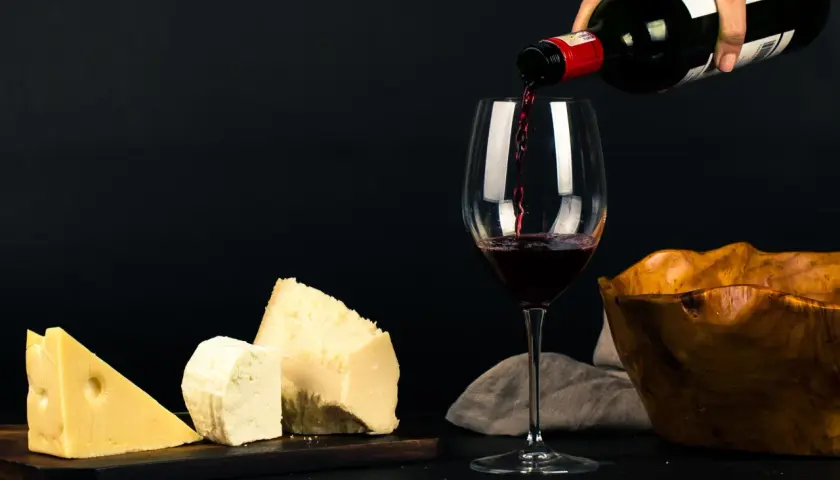 Un verre se remplit de vin rouge, à côté de morceaux de fromage et d'un bol en bois.