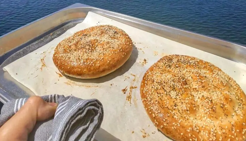 Två gyllene ramazan pidesi, toppade med sesamfrön, vilar på ett bakplåtspapper vid havet.