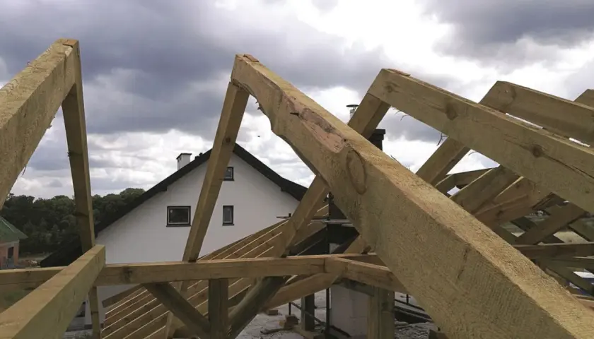 Montaż więźby dachowej w toku. Drewniane belki tworzą konstrukcję dachu nad białym domem z kominem.