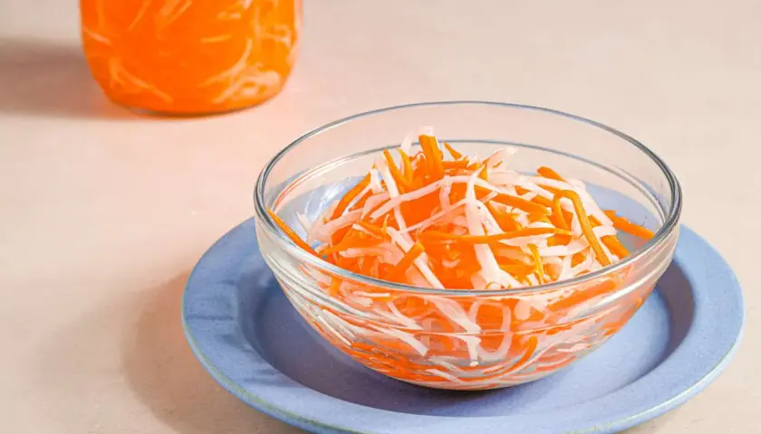 A bowl of vibrant japanese pickled carrots and daikon radish sits on a blue plate, with a jar of the same pickled vegetables in the background.