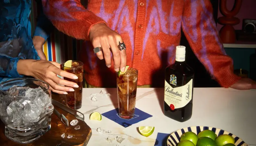 Dwie osoby przygotowują drinki z whisky i colą, z butelką Ballantine's, lodem i limonkami.
