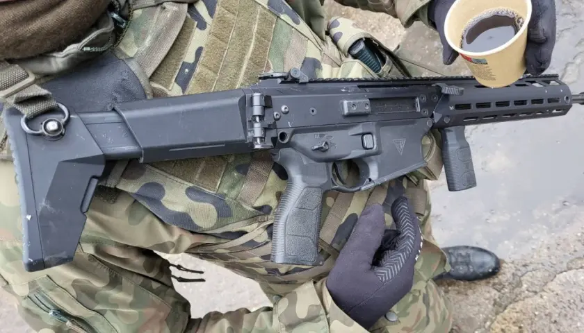 Żołnierz w kamuflażu trzyma karabin i kubek kawy. Czy to broń kolekcjonerska czy sportowa?