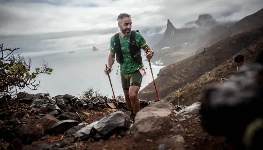 Corredor de montaña con bastones asciende un sendero rocoso, superando un gran desnivel positivo con el mar y acantilados al fondo.