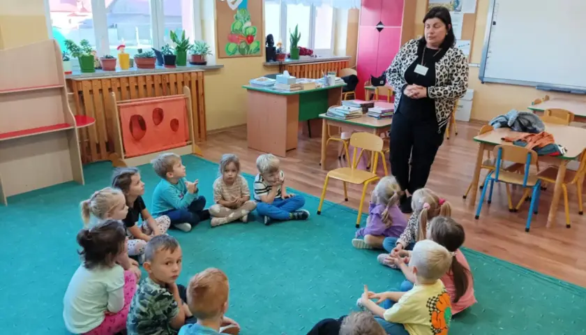Kobieta w panterkowej kurtce stoi przed grupą dzieci siedzących w kręgu na zielonej wykładzinie.