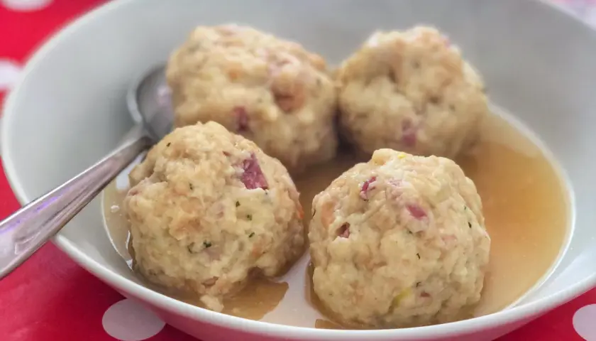 Canederli in brodo, un classico dei piatti tipici Alto Adige, serviti in una ciotola bianca su un tavolo rosso a pois bianchi.
