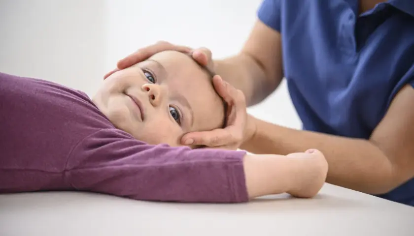 Sanfte Hände einer Osteopathin untersuchen den Kopf eines lächelnden Kleinkindes.