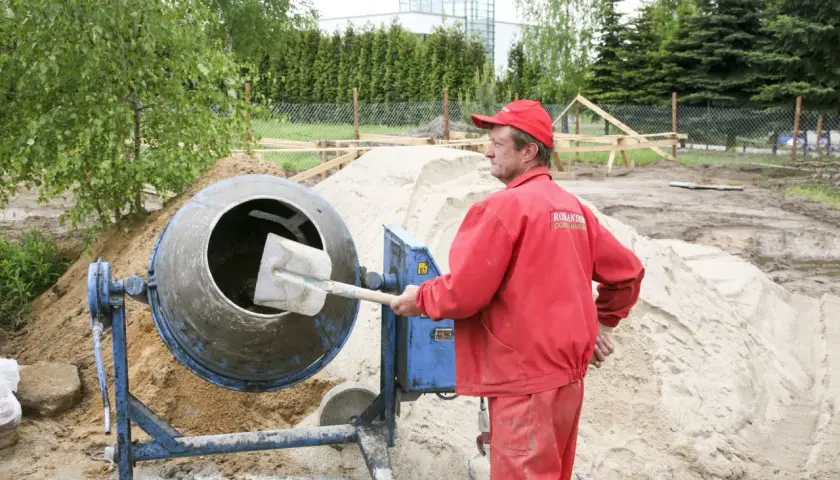 Budowlaniec w czerwonym ubraniu obsługuje betoniarkę na placu budowy, obok sterty piasku.