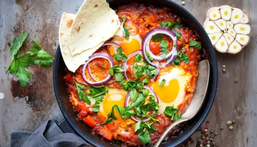 Délicieuse recette de chakchouka dans une poêle, garnie d'œufs, d'oignons rouges et de persil frais. Servi avec du pain pita.