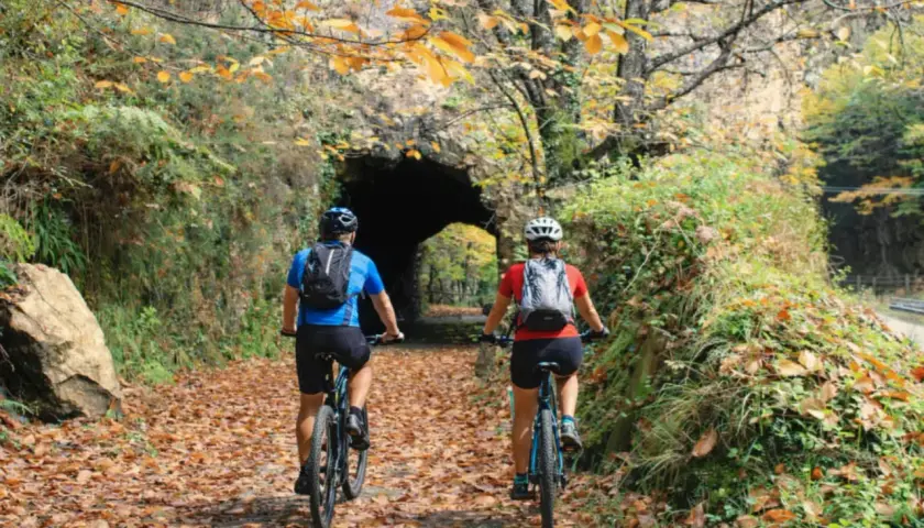 Ciclistas pedalean por la senda del oso, cubierta de hojas otoñales, hacia un túnel de piedra.